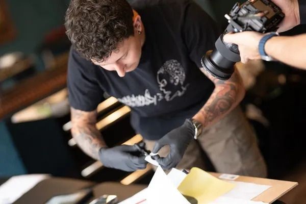 Person with tattoos preparing a print while being photographed, wearing gloves and a black t-shirt.